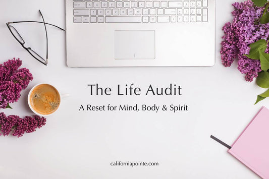 A flatlay image of a white desk, laptop keyboard, pink journal, eyeglasses, tumeric tea in a mug, and purple flowers with 'The Life Audit A Reset for Mind, Body & Spirit' in black text in the center, 'californiapointe.com' in black text on the bottom