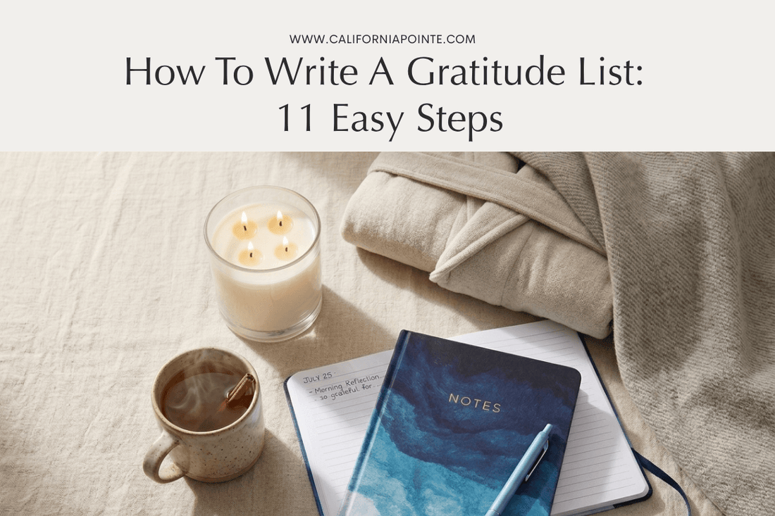 An open blue and teal watercolor design journal on a linen surface with a four-wick lit candle and a ceramic mug with a cinnamon stick to the left, and a folded waffle knit bathrobe and throw blanket to the right.